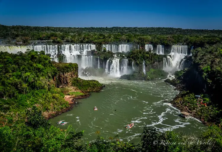 A panoramic view of the Iguazu Falls waterfall system with multiple cascades surrounded by lush greenery. Boats with tourists are visible on the river below. The waterfalls straddle the border of Argentina and Brazil in South America.