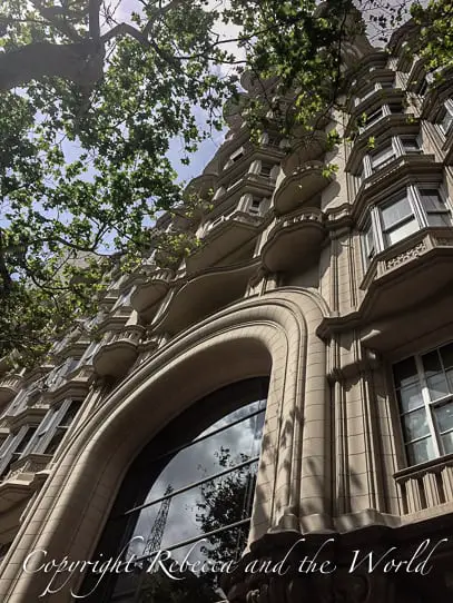 Exterior view of Palacio Barolo showcasing the intricate architecture, featuring rounded balconies with elaborate stonework. The photo is taken from a low angle, highlighting the building's height and the ornate façade against a backdrop of clear sky and tree branches at the top corner.