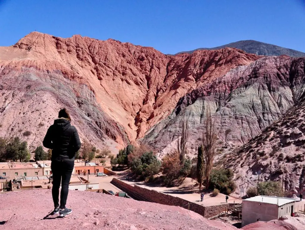 A woman (the author of this article) stands with their back to the camera, overlooking a dramatic landscape of multicolored mountain slopes in shades of red, purple, and gray - this is the Cerro de Siete Colores (Hill of 7 Colours). A small village with terracotta rooftops is nestled at the mountain's base beneath a clear blue sky. The Cerro de Siete Colores in north Argentina is absolutely beautiful - base yourself in Purmamarca to see it before and after sunset when the colours are at their most rich.