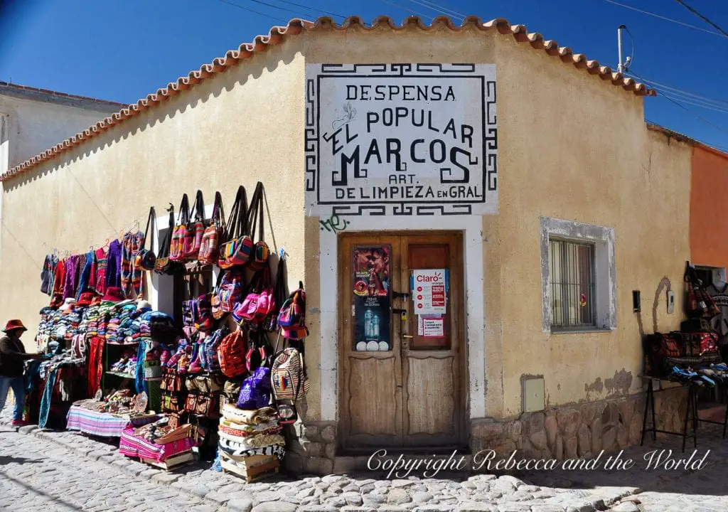 A colorful storefront with traditional textiles and handicrafts displayed outside. The sign above reads 