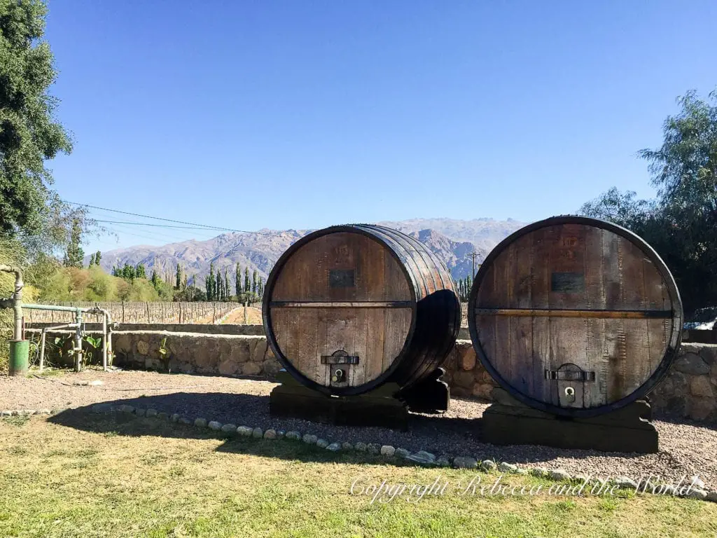 Two large, weathered wine barrels situated outdoors with a backdrop of mountains and a clear sky. The barrels are at a winery in Cafayate, north Argentina.
