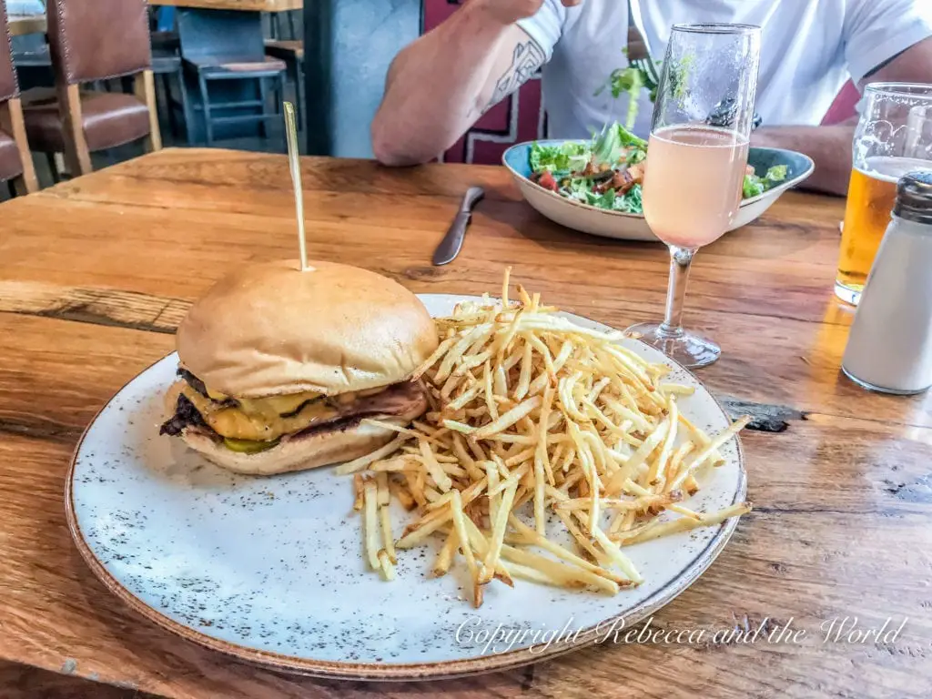 A close-up image of a meal at a restaurant. The main focus is on a plate with a cheeseburger and a large serving of thin, crispy fries. The burger has a toasted bun and visible layers of cheese and pickles. In the background, a person is partially visible, sitting at the table with a salad, a glass of pink beverage, and a glass of beer.