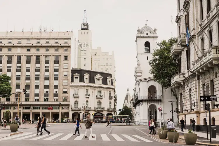 A busy intersection in Buenos Aires, Argentina, with pedestrians crossing, featuring a blend of modern and historic architecture, including a prominent white building with a clock tower.