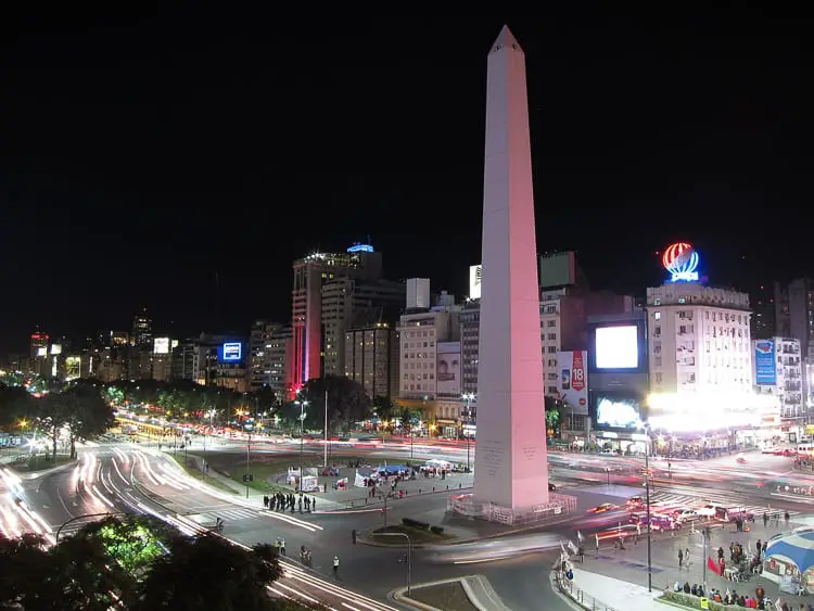 A nighttime cityscape showing a well-lit obelisk monument at the center of a traffic circle, with streaks of light from moving vehicles. The Obelisk in Buenos Aires, Argentina, is one of the most photographed spots in the city.