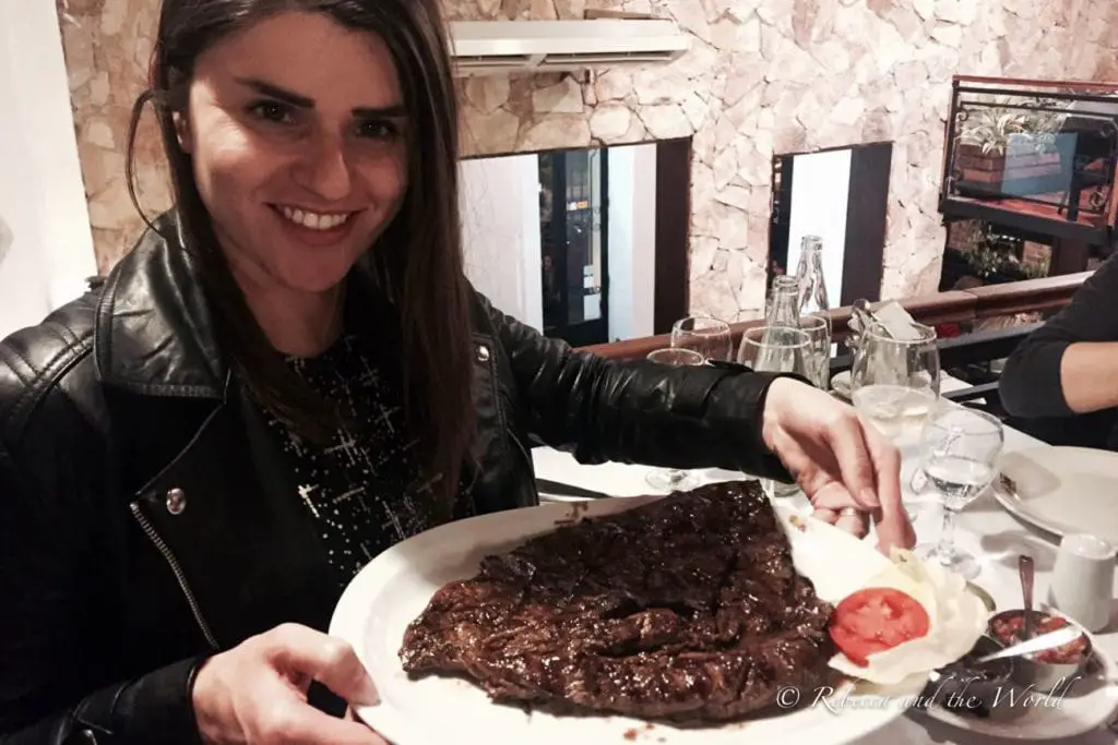 A woman in a leather jacket (the author of the article) holds up a plate with a large piece of cooked steak on it, with a side of tomato slices. 