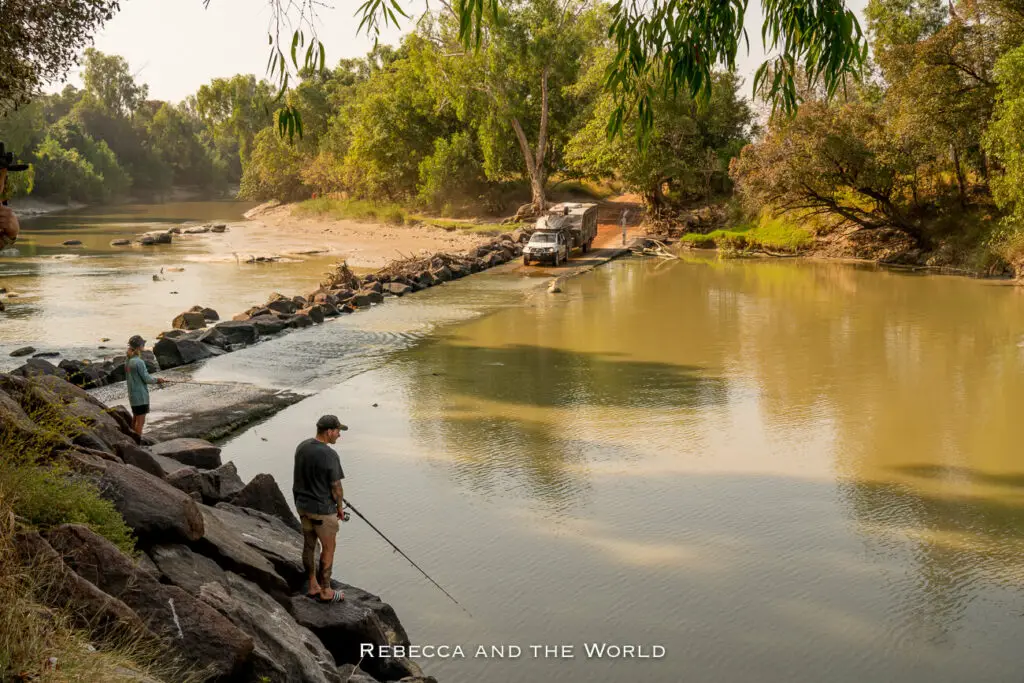 Two individuals fishing from a rocky outcrop by a serene river, with a 4WD vehicle crossing a shallow causeway in the background. This is Cahills Crossing in Kakadu National Park, notorious for being where crocodiles gather at high tide to catch fish.