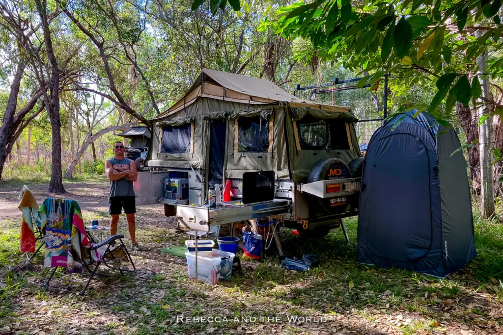 A man - the author's husband - standing beside a camping setup in the woods with a tent mounted on a 4WD vehicle and camping chairs and equipment arranged around. This is Maguk campground in Kakadu National Park.