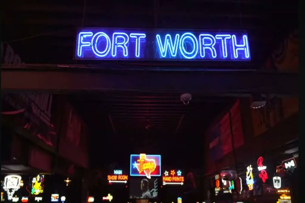 The entrance to a lively venue in Fort Worth, with a prominent neon sign reading "FORT WORTH." Below the sign, there are various other illuminated signs and decorations, suggesting a bustling nightlife scene. The interior is dimly lit, with colourful lights creating a vibrant atmosphere.