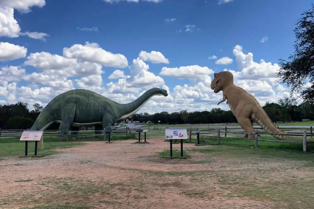 An outdoor scene at a dinosaur park, featuring large statues of a green Brontosaurus and a brown Tyrannosaurus rex. The Brontosaurus is on the left, facing the T-rex on the right. The sky is partly cloudy, and the park has grassy areas and a dirt path, with informational signs in the foreground. Glen Rose is a fun place to visit near Dallas.