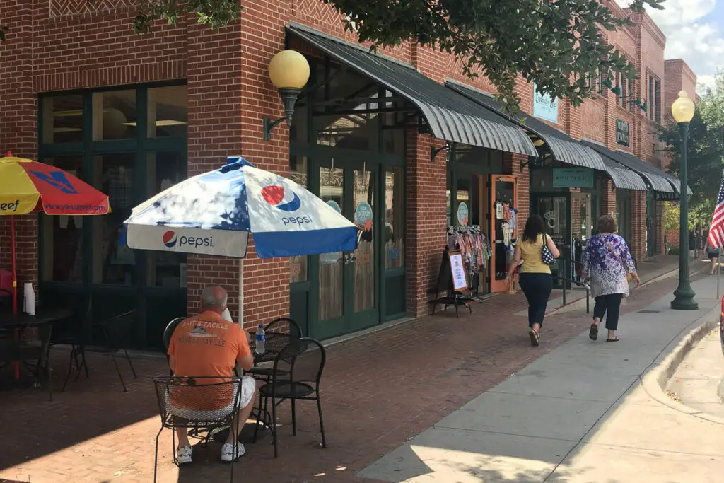A bustling sidewalk scene in the small town of Grapevine near Dallas TX, with a brick building housing various shops. The storefronts have large windows and metal awnings. There are outdoor tables with umbrellas branded with Pepsi logos, where people are seated. Two women are walking down the sidewalk, and an American flag is visible on the right.