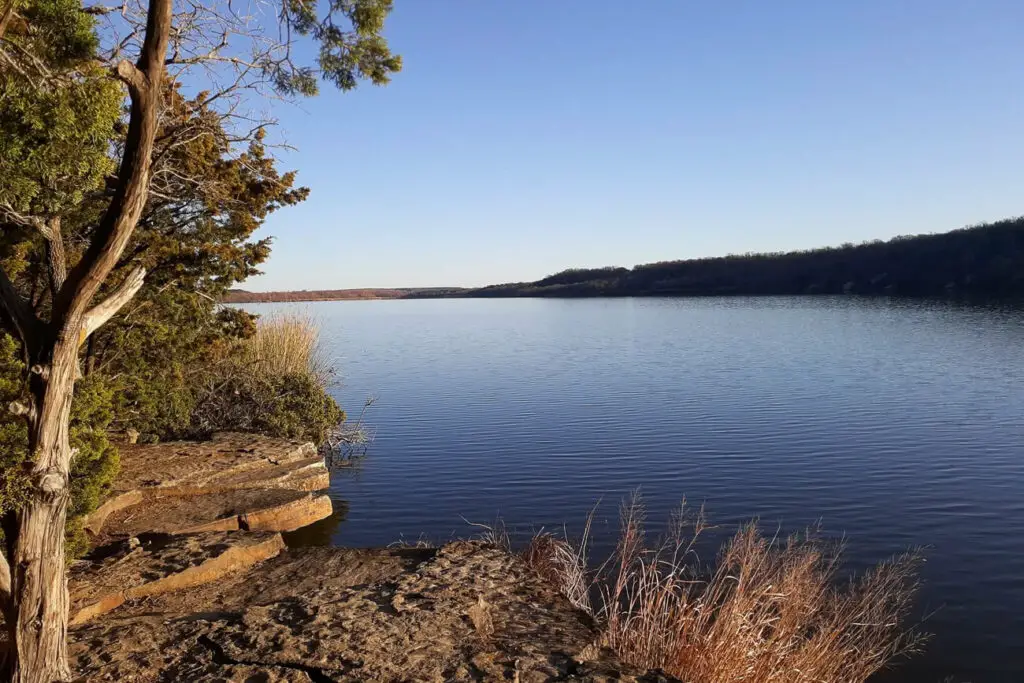 A serene lakeside view featuring a rocky shoreline, sparse vegetation, and clear, calm water extending into the distance. The sky is clear and blue, suggesting a peaceful and pleasant day. Trees and bushes are visible on the left side, with the water reflecting the blue sky and the distant horizon. This is Lake Mineral Wells, a great day trip from Dallas destination.